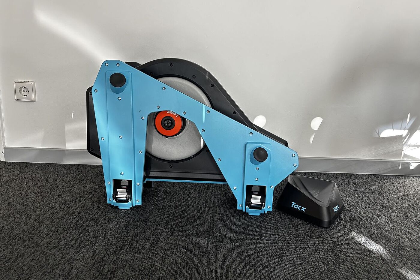 Tacx eingeklappt Ein Smarttrainer mit eingeklappten Beinen steht vor einer weißen Wand.